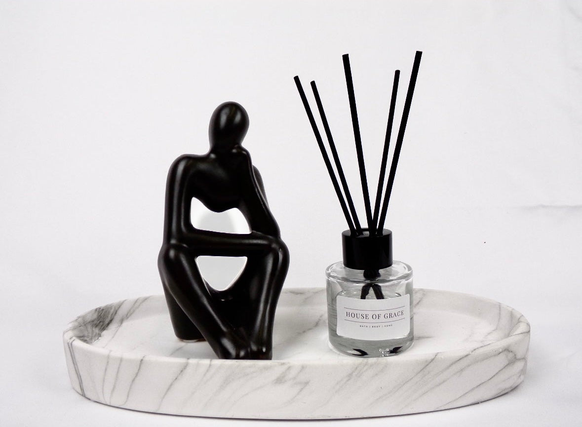 Reed diffuser in sleek clear glass jar with thin black reeds, on a white marble plate, white background, and with a black sculpture of a person sitting next to it.