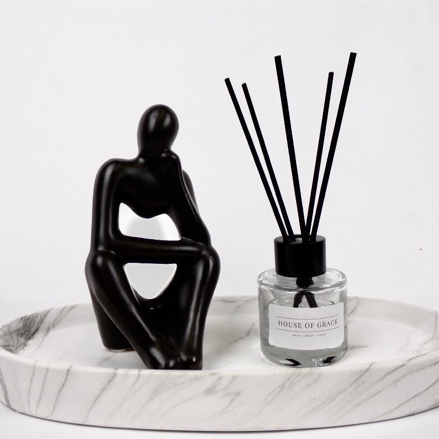 Reed diffuser in sleek clear glass jar with thin black reeds, on a white marble plate, white background, and with a black sculpture of a person sitting next to it.
