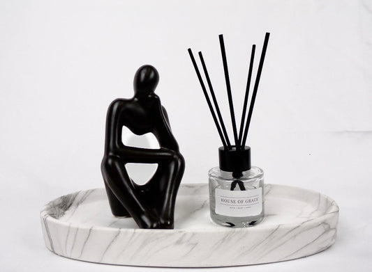 Reed diffuser in sleek clear glass jar with thin black reeds, on a white marble plate, white background, and with a black sculpture of a person sitting next to it.